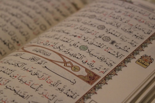 An open Arabic book displaying intricate calligraphy with decorative elements on the margins. The text is organized into lines with a mix of black and red script, and features ornamental designs and symbols.