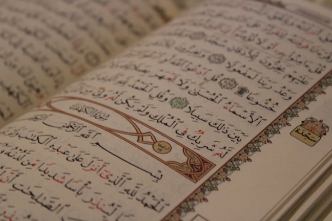An open Arabic book displaying intricate calligraphy with decorative elements on the margins. The text is organized into lines with a mix of black and red script, and features ornamental designs and symbols.