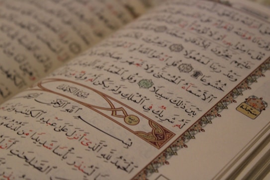 An open Arabic book displaying intricate calligraphy with decorative elements on the margins. The text is organized into lines with a mix of black and red script, and features ornamental designs and symbols.