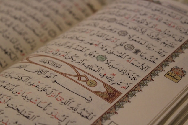 An open Arabic book displaying intricate calligraphy with decorative elements on the margins. The text is organized into lines with a mix of black and red script, and features ornamental designs and symbols.