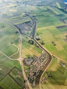 An aerial view of a rural landscape featuring an intricate network of green agricultural fields divided by narrow water channels. A small village with dense housing is nestled in the center, intersected by a meandering river. Roads with a roundabout can be seen connecting different parts of the area, providing a sense of connectivity.