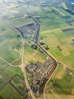 An aerial view of a rural landscape featuring an intricate network of green agricultural fields divided by narrow water channels. A small village with dense housing is nestled in the center, intersected by a meandering river. Roads with a roundabout can be seen connecting different parts of the area, providing a sense of connectivity.