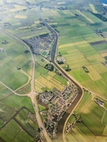 An aerial view of a rural landscape featuring an intricate network of green agricultural fields divided by narrow water channels. A small village with dense housing is nestled in the center, intersected by a meandering river. Roads with a roundabout can be seen connecting different parts of the area, providing a sense of connectivity.