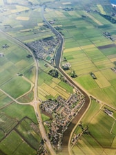 An aerial view of a rural landscape featuring an intricate network of green agricultural fields divided by narrow water channels. A small village with dense housing is nestled in the center, intersected by a meandering river. Roads with a roundabout can be seen connecting different parts of the area, providing a sense of connectivity.