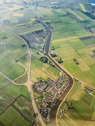 An aerial view of a rural landscape featuring an intricate network of green agricultural fields divided by narrow water channels. A small village with dense housing is nestled in the center, intersected by a meandering river. Roads with a roundabout can be seen connecting different parts of the area, providing a sense of connectivity.