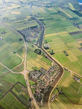 An aerial view of a rural landscape featuring an intricate network of green agricultural fields divided by narrow water channels. A small village with dense housing is nestled in the center, intersected by a meandering river. Roads with a roundabout can be seen connecting different parts of the area, providing a sense of connectivity.