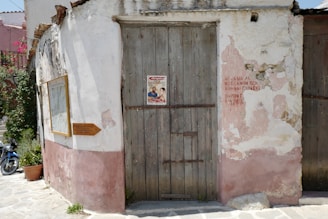 An old, worn building wall with a weathered wooden door and faded pink and white plaster. A poster is pinned to the door, and Greek writing is visible nearby on the wall. A framed sign and a small wooden directional sign are attached to the wall. Potted plants and a blue motorbike can be seen on the paved street.
