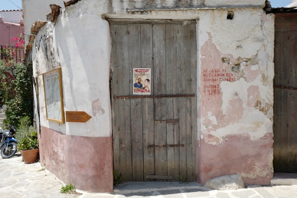 An old, worn building wall with a weathered wooden door and faded pink and white plaster. A poster is pinned to the door, and Greek writing is visible nearby on the wall. A framed sign and a small wooden directional sign are attached to the wall. Potted plants and a blue motorbike can be seen on the paved street.