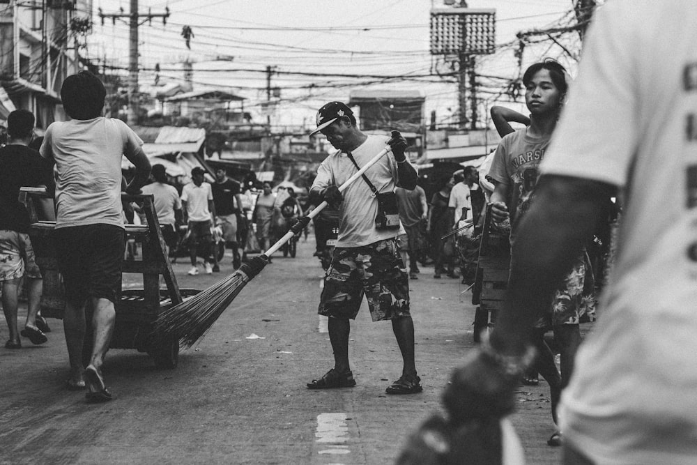 grayscale photography of man sweeping on pavement by person pushing kart