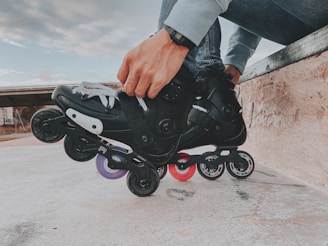 A person is tightening the straps on black rollerblades while sitting on a concrete ledge. The rollerblades have multiple wheels in different colors, including purple and red. The person is wearing a watch and casual clothing.