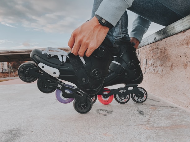 A person is tightening the straps on black rollerblades while sitting on a concrete ledge. The rollerblades have multiple wheels in different colors, including purple and red. The person is wearing a watch and casual clothing.