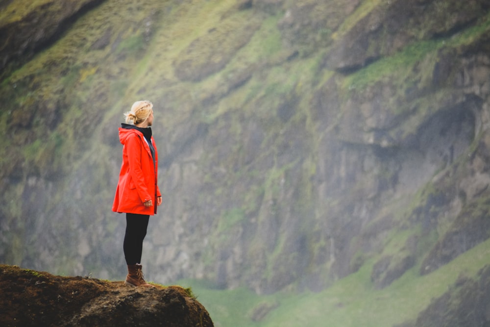 Frau im orangefarbenen Parka steht auf einem Felsen mit Blick auf die Klippe