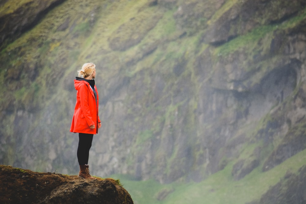 mulher em parka laranja de pé na rocha de frente para o penhasco