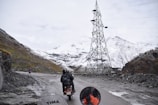 A mountainous landscape with a snow-covered terrain is visible. A motorcyclist, wearing a helmet and backpack, rides along a narrow, winding road. The road is bordered by rocky cliffs, and a tall electrical tower stands to the right. A few other motorcyclists can be seen in the distance. The sky is overcast, adding a sense of chilliness to the scene.