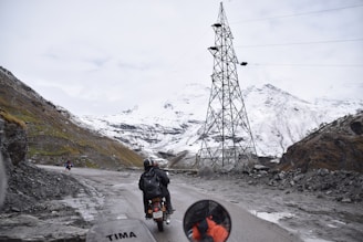 A mountainous landscape with a snow-covered terrain is visible. A motorcyclist, wearing a helmet and backpack, rides along a narrow, winding road. The road is bordered by rocky cliffs, and a tall electrical tower stands to the right. A few other motorcyclists can be seen in the distance. The sky is overcast, adding a sense of chilliness to the scene.