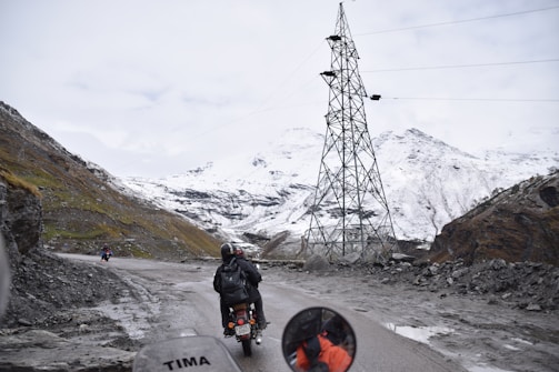A mountainous landscape with a snow-covered terrain is visible. A motorcyclist, wearing a helmet and backpack, rides along a narrow, winding road. The road is bordered by rocky cliffs, and a tall electrical tower stands to the right. A few other motorcyclists can be seen in the distance. The sky is overcast, adding a sense of chilliness to the scene.