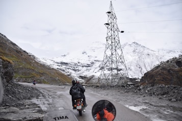 A mountainous landscape with a snow-covered terrain is visible. A motorcyclist, wearing a helmet and backpack, rides along a narrow, winding road. The road is bordered by rocky cliffs, and a tall electrical tower stands to the right. A few other motorcyclists can be seen in the distance. The sky is overcast, adding a sense of chilliness to the scene.