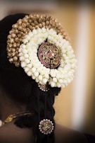 A woman's hair is adorned with intricate floral garlands and decorative hair accessories. The floral arrangement consists of white jasmine flowers and golden buds, with a central decorative piece featuring red and green stones. Additional smaller accessories are integrated into the braid.