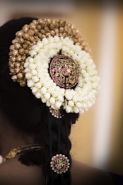 A woman's hair is adorned with intricate floral garlands and decorative hair accessories. The floral arrangement consists of white jasmine flowers and golden buds, with a central decorative piece featuring red and green stones. Additional smaller accessories are integrated into the braid.