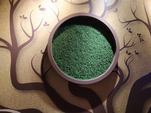 A circular wall-mounted planter filled with green moss or grass is set against a textured wall with artistic tree and leaf patterns. The artwork has abstract, flowing branches extending across the wall, creating a natural and organic aesthetic.