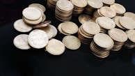 A collection of stacks and scattered coins on a dark surface. The coins are circular and metallic, with some displaying the denomination and an emblem. The central focus is on a group of Indian five-rupee coins.
