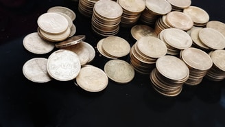 A collection of stacks and scattered coins on a dark surface. The coins are circular and metallic, with some displaying the denomination and an emblem. The central focus is on a group of Indian five-rupee coins.