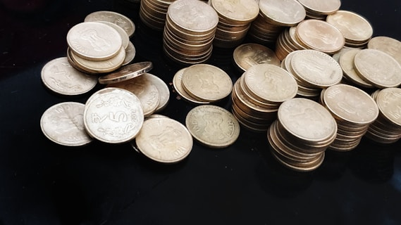 A collection of stacks and scattered coins on a dark surface. The coins are circular and metallic, with some displaying the denomination and an emblem. The central focus is on a group of Indian five-rupee coins.