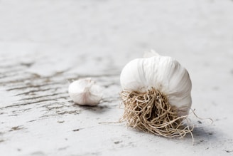 A close-up of a bulb of garlic resting on a textured, light-colored wooden surface. The garlic's roots are visible, and there is another smaller clove in the background.