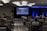 A conference room filled with attendees seated in rows facing a large screen displaying '#PRODUCTCON Los Angeles' along with sponsor logos. The room is dimly lit with bright blue accent lighting on the walls and ceiling, and people appear to be engaged and attentive.