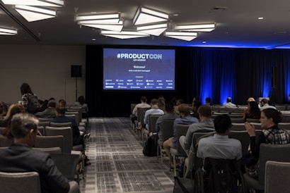 A conference room filled with attendees seated in rows facing a large screen displaying '#PRODUCTCON Los Angeles' along with sponsor logos. The room is dimly lit with bright blue accent lighting on the walls and ceiling, and people appear to be engaged and attentive.