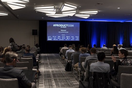 A conference room filled with attendees seated in rows facing a large screen displaying '#PRODUCTCON Los Angeles' along with sponsor logos. The room is dimly lit with bright blue accent lighting on the walls and ceiling, and people appear to be engaged and attentive.