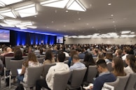 A large audience is seated in a conference room, focusing on a speaker on stage. The room is well-lit with modern ceiling lights, and two large screens flank the stage displaying visual content. Most people are facing forward with several of them taking notes or using laptops.