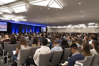 A large audience is seated in a conference room, focusing on a speaker on stage. The room is well-lit with modern ceiling lights, and two large screens flank the stage displaying visual content. Most people are facing forward with several of them taking notes or using laptops.