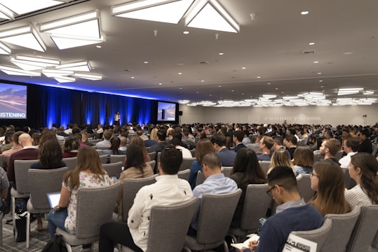A large audience is seated in a conference room, focusing on a speaker on stage. The room is well-lit with modern ceiling lights, and two large screens flank the stage displaying visual content. Most people are facing forward with several of them taking notes or using laptops.