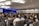 A large audience is seated in a conference room, focusing on a speaker on stage. The room is well-lit with modern ceiling lights, and two large screens flank the stage displaying visual content. Most people are facing forward with several of them taking notes or using laptops.
