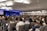 A large audience is seated in a conference room, focusing on a speaker on stage. The room is well-lit with modern ceiling lights, and two large screens flank the stage displaying visual content. Most people are facing forward with several of them taking notes or using laptops.