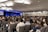 A large audience is seated in a conference room, focusing on a speaker on stage. The room is well-lit with modern ceiling lights, and two large screens flank the stage displaying visual content. Most people are facing forward with several of them taking notes or using laptops.