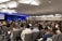 A large audience is seated in a conference room, focusing on a speaker on stage. The room is well-lit with modern ceiling lights, and two large screens flank the stage displaying visual content. Most people are facing forward with several of them taking notes or using laptops.