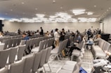 A large conference room with numerous rows of grey chairs, partially filled with people. The room has a modern ceiling design with distinctive lighting fixtures. Attendees are engaged in conversations or looking at their electronic devices. The atmosphere suggests a formal or professional event.