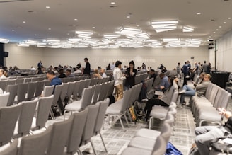 A large conference room with numerous rows of grey chairs, partially filled with people. The room has a modern ceiling design with distinctive lighting fixtures. Attendees are engaged in conversations or looking at their electronic devices. The atmosphere suggests a formal or professional event.