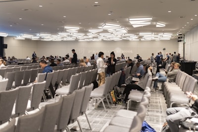 A large conference room with numerous rows of grey chairs, partially filled with people. The room has a modern ceiling design with distinctive lighting fixtures. Attendees are engaged in conversations or looking at their electronic devices. The atmosphere suggests a formal or professional event.