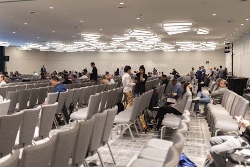 A large conference room with numerous rows of grey chairs, partially filled with people. The room has a modern ceiling design with distinctive lighting fixtures. Attendees are engaged in conversations or looking at their electronic devices. The atmosphere suggests a formal or professional event.