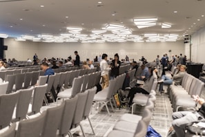 A large conference room with numerous rows of grey chairs, partially filled with people. The room has a modern ceiling design with distinctive lighting fixtures. Attendees are engaged in conversations or looking at their electronic devices. The atmosphere suggests a formal or professional event.