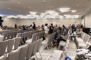 A large conference room with numerous rows of grey chairs, partially filled with people. The room has a modern ceiling design with distinctive lighting fixtures. Attendees are engaged in conversations or looking at their electronic devices. The atmosphere suggests a formal or professional event.
