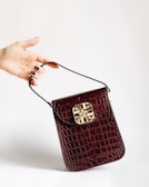 A hand with painted nails holds a small, dark red crocodile leather handbag with a gold decorative clasp. The background is plain and light-colored, accentuating the bag and hand.