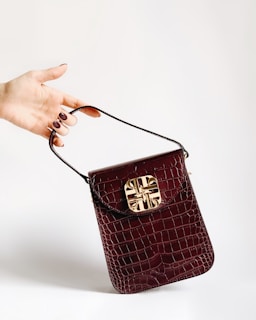 A hand with painted nails holds a small, dark red crocodile leather handbag with a gold decorative clasp. The background is plain and light-colored, accentuating the bag and hand.