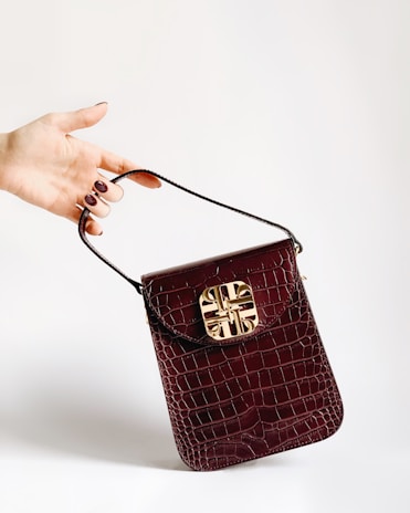 A hand with painted nails holds a small, dark red crocodile leather handbag with a gold decorative clasp. The background is plain and light-colored, accentuating the bag and hand.