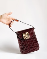 A hand with painted nails holds a small, dark red crocodile leather handbag with a gold decorative clasp. The background is plain and light-colored, accentuating the bag and hand.