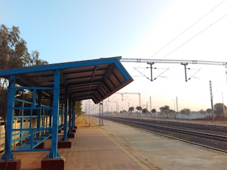 A small, empty train platform with a blue metal and wooden shelter on the left side. The platform is lined with a fence and bordered by railway tracks on the right. Overhead power lines run above the tracks. Trees and distant shrubs are visible in the background under a clear sky.