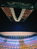 A futuristic architectural structure with illuminated patterns. The design features symmetrical lines and vibrant lighting that create a captivating, modern aesthetic. The reflection enhances the visual impact, lending a sense of depth and symmetry to the scene.
